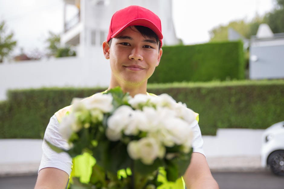 A young male delivery person with light skin, dark hair, and a friendly expression, wearing a red cap and a reflective yellow vest, holds a bouquet of fresh white flowers with green foliage towards th