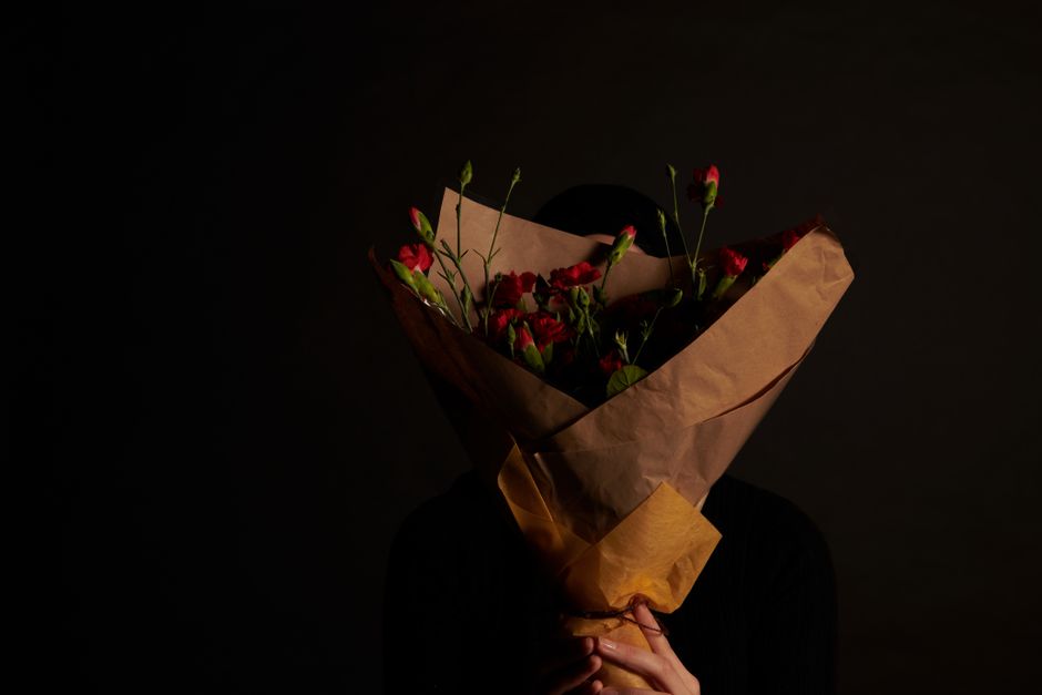 A person holding a bouquet of fresh red and pink flowers wrapped in brown kraft paper and tied with a pink ribbon, partially covering their face. The bouquet features tightly clustered blooms with gre