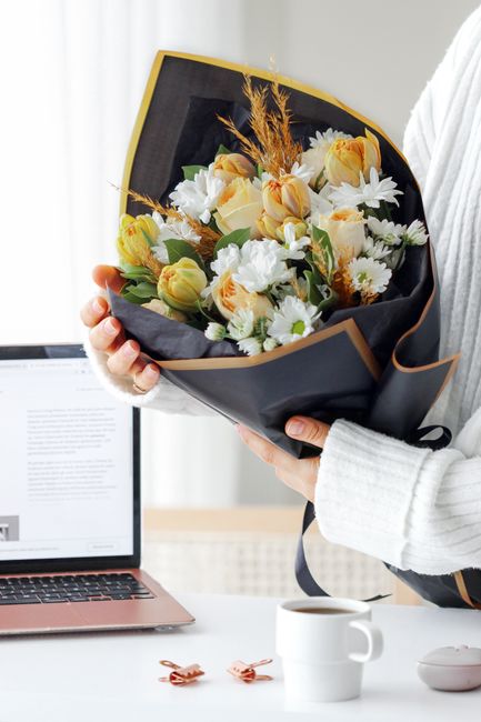 A person dressed in a white sweater is holding a bouquet of fresh flowers, featuring white daisies, creamy yellow roses, and delicate white blossoms, arranged with orange wheat-like stalks. The bouque