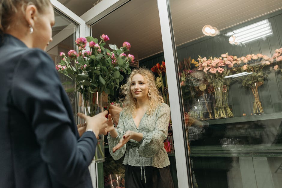 A woman with curly blonde hair, dressed in a light-colored floral blouse, is receiving a bouquet of pink roses wrapped in brown paper from a florist dressed in a dark jacket. The florist's hand is gen