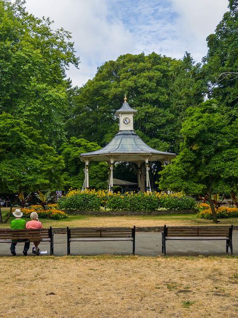 A white, Victorian-style bandstand with a clock on its upper section, surrounded by lush green trees and vibrant yellow flowerbeds in Gladstone Park. In the foreground, two elderly people sit on a woo