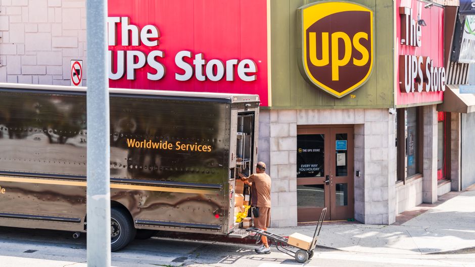 A delivery person in casual clothing is unloading a box from a large UPS freight truck parked outside The UPS Store, which has a pink and white facade with the logo prominently displayed. The scene sh