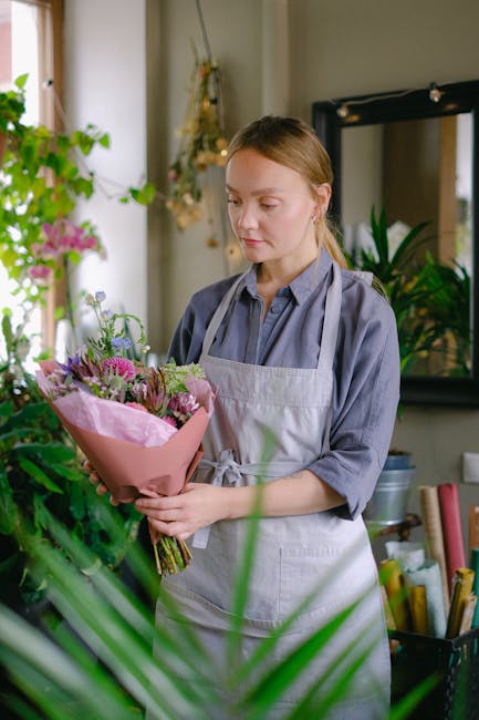 A young woman with light brown hair tied back, wearing a light blue shirt and a white apron, is holding a bouquet of fresh flowers wrapped in pink paper, featuring purple, pink, and green blooms, in a