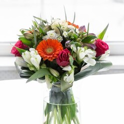 Showcasing a masterful blend of colour and texture, this abundant floral ensemble captures the uplifting energy of Dollis Hill on a sun-dappled spring morning. Creamy and peach gerberas create a gentle halo around the arrangement, their softly arching petals embracing vivid orange centres that radiate warmth. Deep magenta roses, tightly wound and impossibly elegant, add a sense of richness and romantic depth, their hues reminiscent of blossoms found in neighbourhood gardens. White alstroemeria and petite spray roses bring brightness, their multi-petaled forms mingling with hints of pale pink blooms and tiny white filler flowers for a garden-fresh, organic feel. All are held in the gentle grasp of glossy, robust aspidistra leaves, which curve and wrap about the bouquet's base, offering stability and structure akin to the sturdy London plane trees lining Dollis Hill's avenues. Slender leafy accents peek out, lending contrast and an air of effortless abundance. Arranged in a tall, clear glass vase, the bouquet celebrates both craftsmanship and natural vitality, while the soft background light enhances each hue and detail. There's a sense of quiet optimism here-a bouquet as perfect for a convivial brunch as for acknowledging new beginnings along Chapter Road or Church Lane.